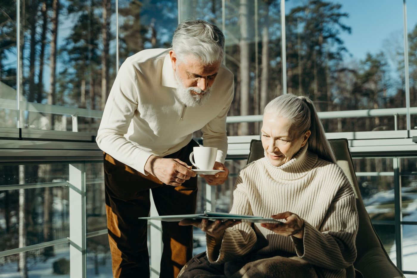 Older woman reading on a sunlit porch while a man carries two mugs toward her