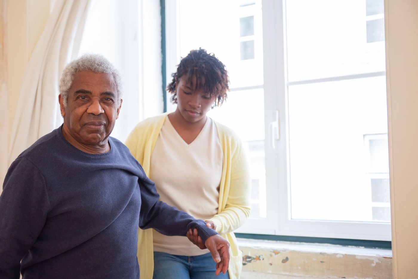Woman supporting an older man by the arm beside a bright window
